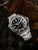 Herrenarmbanduhr mit schwarzem Zifferblatt, silbernem Metallband und orangefarbenen Zeigern