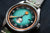 Montre homme en gros plan : cadran turquoise, index orange, bracelet en cuir, couronne