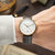 Montre pour homme au poignet, analogique : cadran blanc, aiguilles dorées, bracelet en cuir marron.