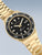 Goldfarbene Herrenuhr mit schwarzem Zifferblatt, Leuchtmarken und Datumsanzeige