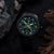 Herrenarmbanduhr mit schwarzem Metallband, grün leuchtende Ziffern und gelbe Zeiger