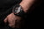 Au poignet : montre homme avec cadran noir et bracelet noir perforé