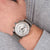 Silberne Herrenarmbanduhr am Handgelenk mit Metall-Mesharmband und weißem Zifferblatt