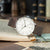 Montre homme avec bracelet en cuir marron, boîtier argenté et cadran blanc sur un livre.