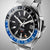 Montre homme en gros plan : cadran noir, lunette bleue, index argentés