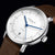 Montre homme, gros plan : cadran blanc avec aiguilles des heures et des minutes bleues, bracelet en cuir marron