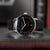 Montre homme avec cadran noir, aiguille des secondes rouge et bracelet en cuir noir.
