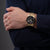 Herrenarmbanduhr am Handgelenk, goldfarbenes Metallband, schwarzes Zifferblatt mit Datum.
