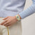 Beige Armbanduhr am Handgelenk, rundes Zifferblatt mit lila Zeigern, beiges Band