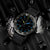 Montre homme avec boîtier gris et cadran noir, index luminescents bleus et verts.