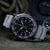 Montre homme avec bracelet textile gris, cadran sombre avec points lumineux et aiguilles orange.