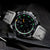 Montre homme avec cadran noir, boîtier argenté et bracelet en cuir noir.