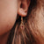 Puce d'oreille dorée avec pierre sombre dans un cadre carré, visible sur l'oreille.