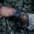 Montre pour homme au poignet en tenue de camouflage : cadran bleu, bracelet foncé, index lumineux visibles.