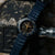 Montre pour homme au poignet sur fond sombre : bracelet bleu, index lumineux dorés sur le cadran.