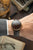 Herrenarmbanduhr am Handgelenk: dunkles Zifferblatt, goldene Lünette, dunkelgraues Lederband