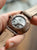 Fond de montre pour homme avec mouvement apparent dans un boîtier gris, bracelet en cuir marron.