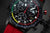 Montre homme, gros plan : cadran noir avec accents rouges et boutons chronographe