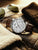 Herrenuhr mit silbernem Zifferblatt und braunem Lederarmband auf brauner Jacke